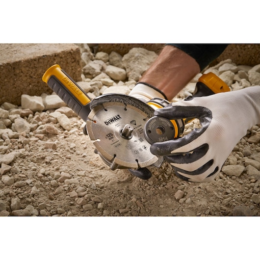 Gloved hands hold a DEWALT DT3559 angle grinder with diamond blade on a rocky surface outdoors.