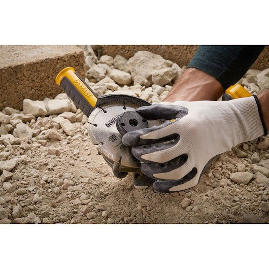 Gloved hand adjusting a DEWALT angle grinder on rocky ground outdoors.