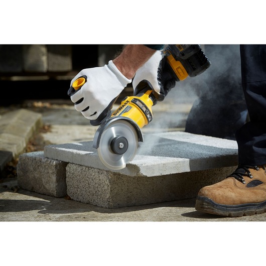 Person uses DEWALT angle grinder DT3711 to cut concrete block outdoors, wearing gloves and safety shoes.