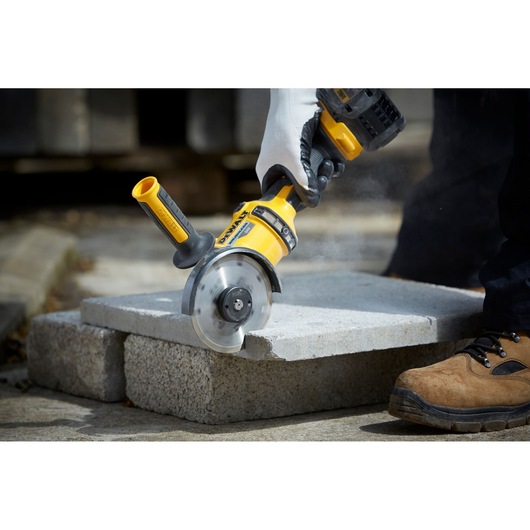 DEWALT grinder cutting concrete slab outdoors, operated by gloved hand and worker in brown safety boots.