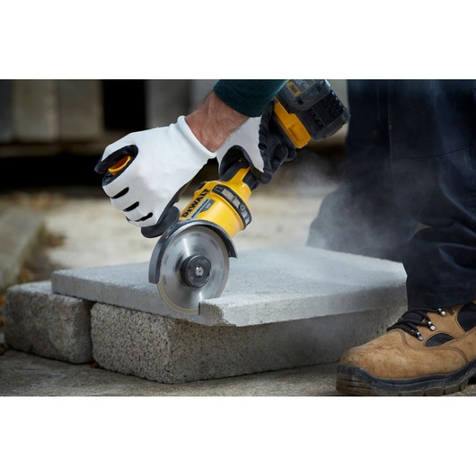 A gloved hand uses a DEWALT tool to cut a concrete slab, with dust rising during the process.