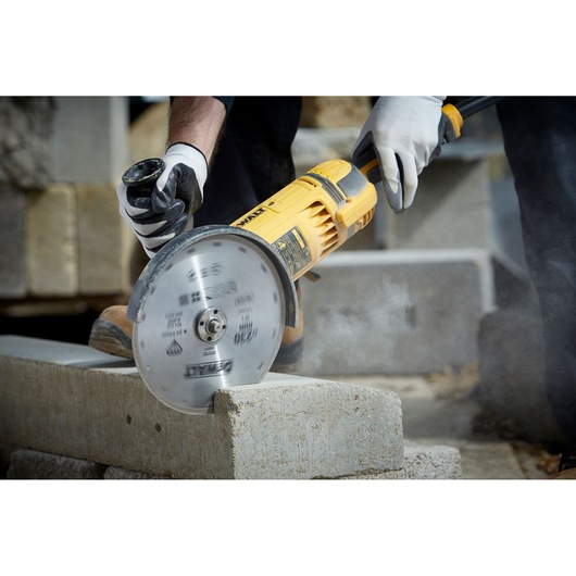 Person uses a DEWALT angle grinder with a diamond blade to cut a concrete block outdoors, wearing safety gloves.