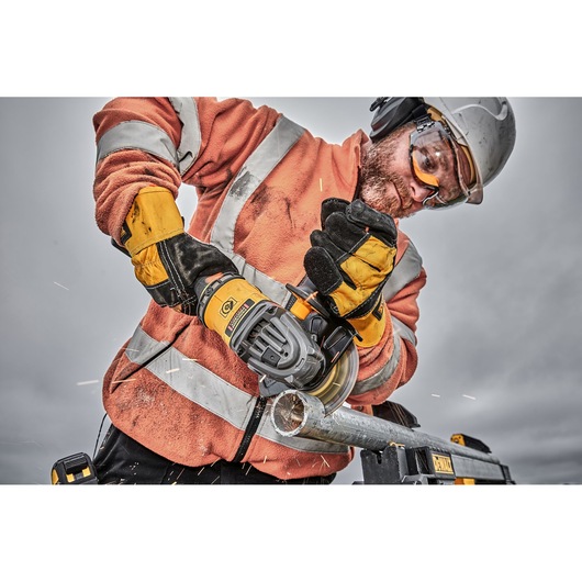 Worker using a DEWALT angle grinder to cut a metal pipe, wearing safety gear and gloves.