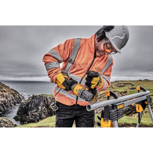 Worker using DEWALT tool to cut a metal pipe outdoors, wearing protective gear and orange safety jacket.
