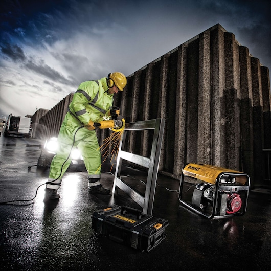 Worker using DEWALT grinder on metal frame outdoors, with DEWALT generator and tool case nearby.
