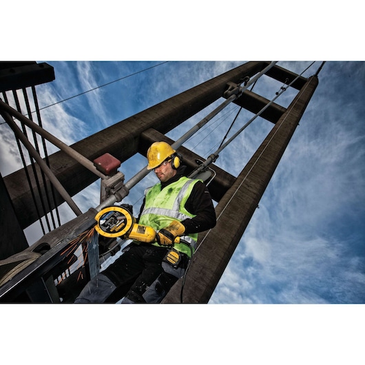 Worker uses DEWALT power tool to cut metal on a bridge, wearing safety gear and helmet under blue sky.