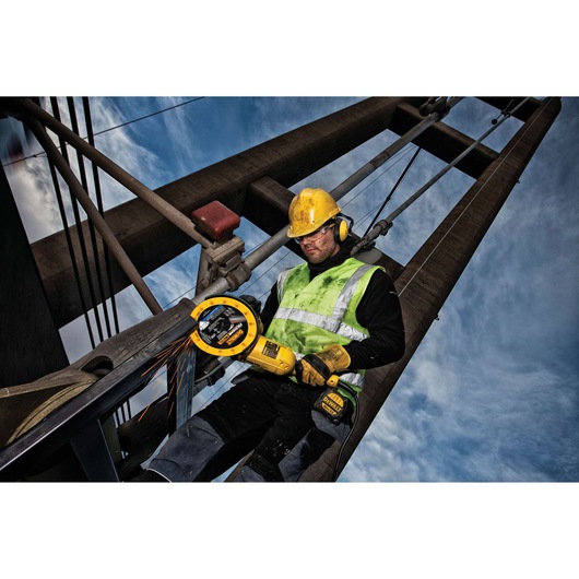 Worker uses DEWALT angle grinder on metal structure outdoors, wearing safety gear and high-vis vest.