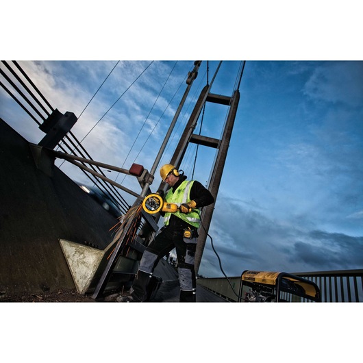 Worker uses a DEWALT grinder on metal near a bridge, wearing safety gear and high-visibility vest.