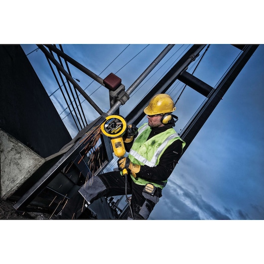 Worker uses DEWALT grinder on metal at construction site, wearing safety gear and high-vis vest.
