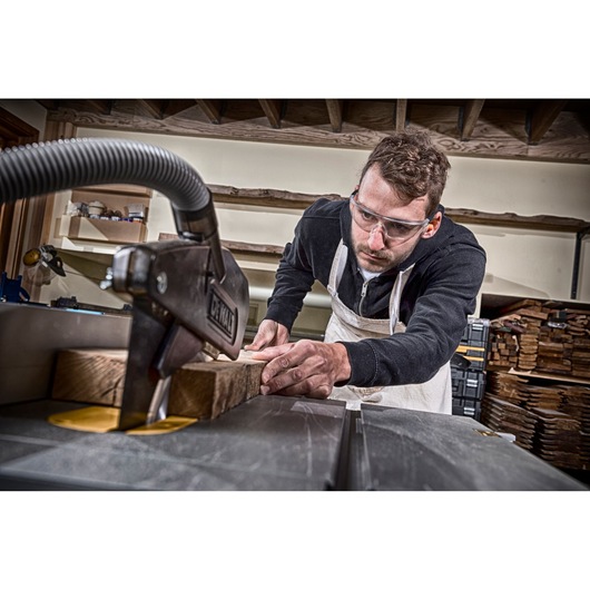 Person cutting wood with DEWALT saw in workshop, wearing apron and focused on woodworking task.