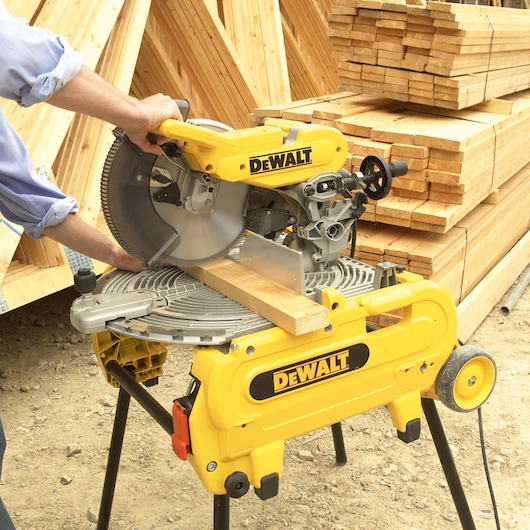 A DEWALT miter saw cutting a piece of wood outdoors, with stacks of lumber in the background. The saw is mounted on a portable stand and operated by a person.