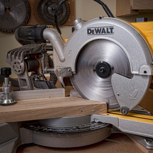 DEWALT miter saw cutting a piece of wood in a workshop, blade spinning, dust visible.
