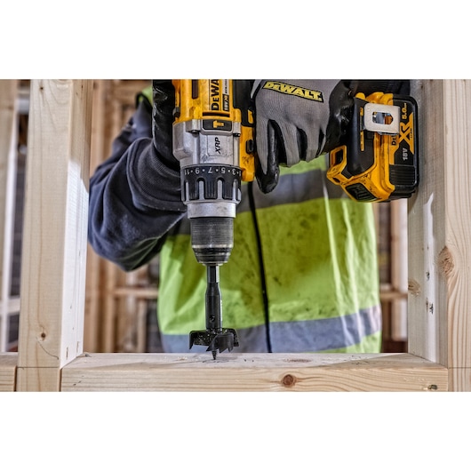 Person uses DEWALT drill to make a hole in wooden frame at construction site, wearing gloves and safety jacket.