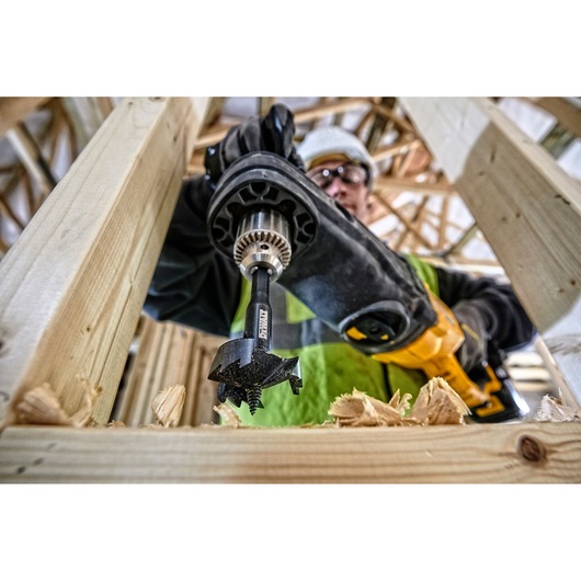 DEWALT drill with DT4582 bit drilling through wood in a construction setting, wood chips visible.