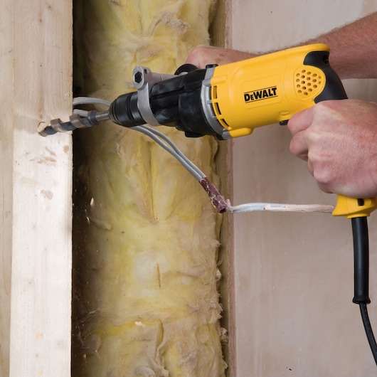 A DEWALT drill is used to bore a hole through wood in a construction setting, with electrical wires visible.