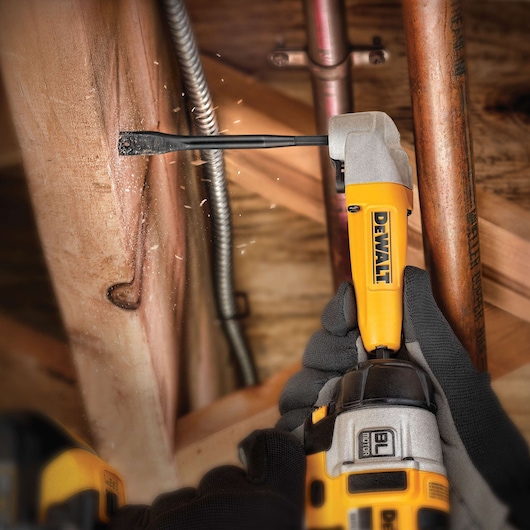 DEWALT drill in use on wooden beam, gloved hands holding, wood chips flying, pipes visible in background.