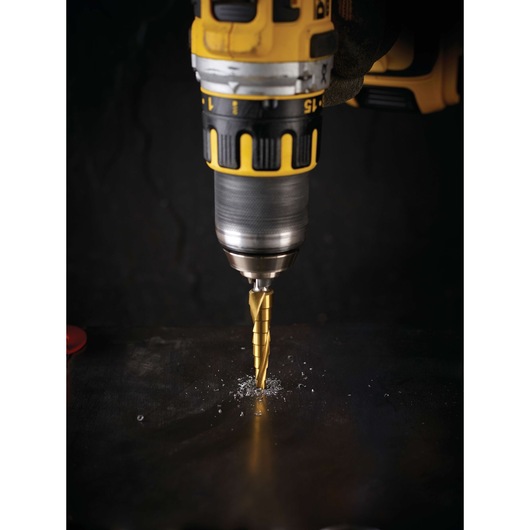 A DEWALT drill with a stepped drill bit is drilling into a dark metal surface, producing metal shavings.