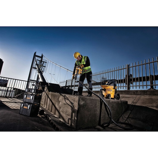Worker uses DEWALT power tool and vacuum on concrete outdoors near a bridge, wearing safety gear and high-visibility vest.