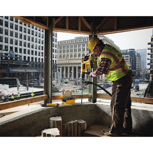 Worker uses DEWALT drill at construction site in city, wearing safety gear and helmet.