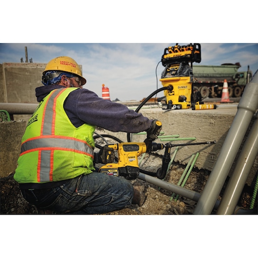 Worker in safety gear uses a DEWALT drill on concrete at a construction site with tools and pipes nearby.