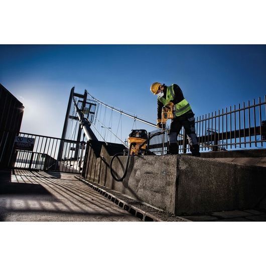 Worker uses DEWALT power tool and vacuum on concrete near a bridge, wearing safety gear and high-visibility vest.