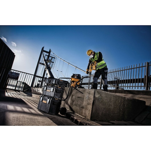 Construction worker using a DEWALT tool near stacked toolboxes on an outdoor site with a bridge in the background.