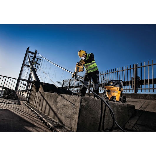 Worker uses DEWALT drill on concrete outdoors by a bridge, wearing safety gear and high-visibility vest.