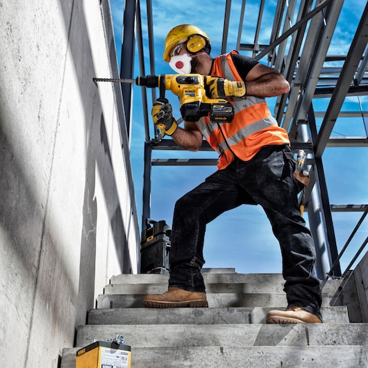 Worker uses DEWALT drill on concrete wall. SKUs: DT60814, DT60814-QZ. Construction site, safety gear visible.