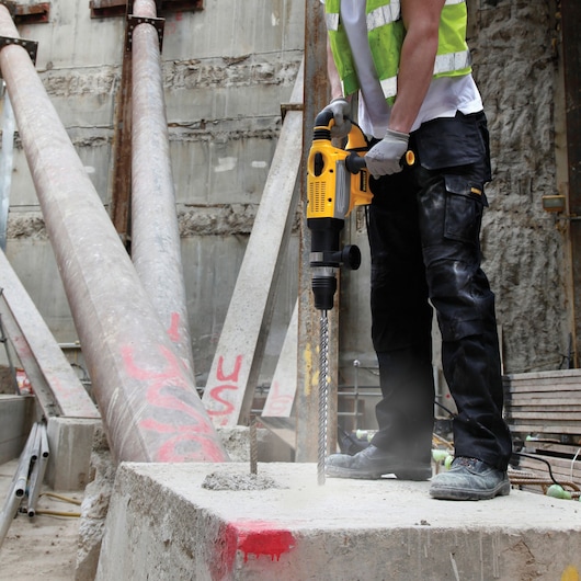 Worker uses a DEWALT hammer drill on concrete at a construction site, wearing safety gear and gloves.