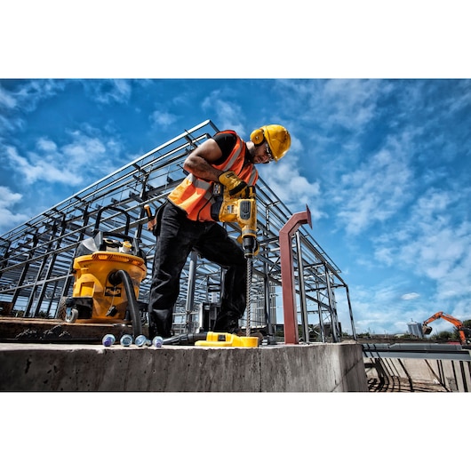 Worker using DEWALT drill at construction site with steel structure, yellow tools, and clear blue sky.