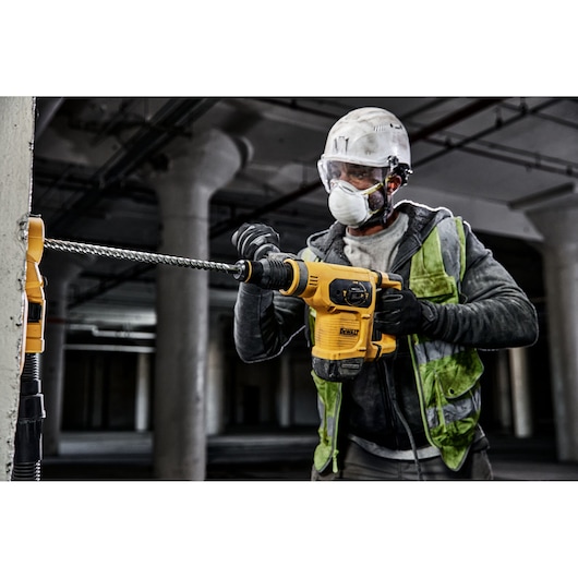 Worker uses a DEWALT rotary hammer drill on a concrete wall in an industrial setting, wearing safety gear.