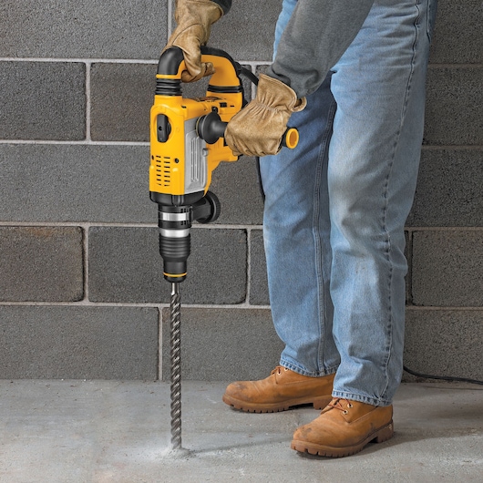 Person uses yellow DEWALT rotary hammer drill with DT60822 bit to drill into concrete floor near a brick wall.