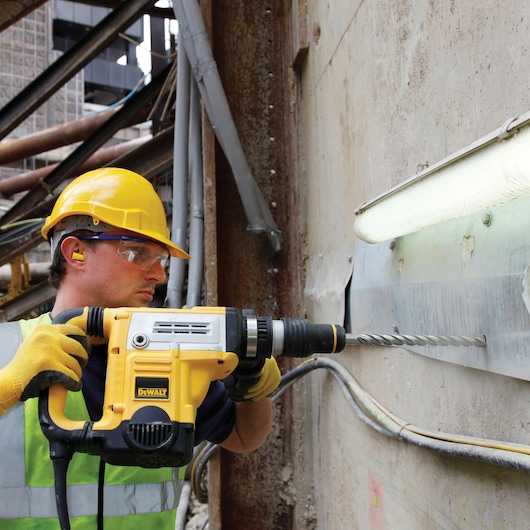 Worker uses DEWALT rotary hammer drill on a concrete wall at construction site, wearing safety gear and helmet.