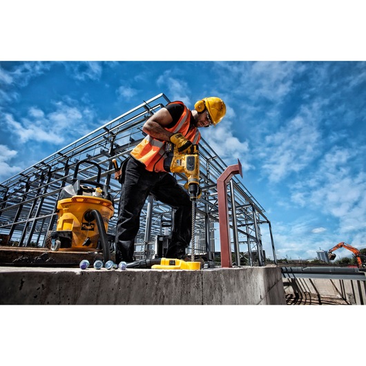 Worker using a DEWALT drill at a construction site with steel structure and equipment under a blue sky.