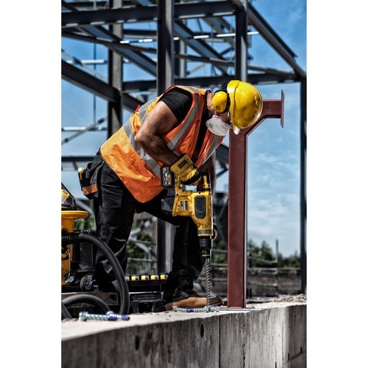 Worker uses DEWALT drill on concrete at construction site, wearing safety gear and orange vest.