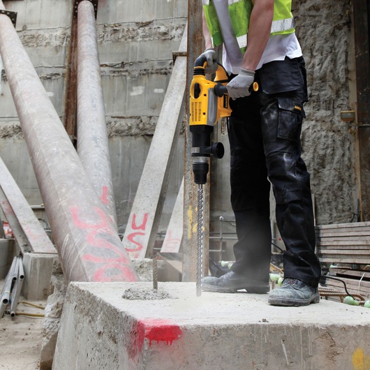 Worker using yellow DEWALT drill DT60823 to drill into concrete at construction site with safety gear.