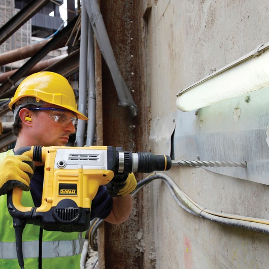 Worker uses a DEWALT drill to make a hole in a concrete wall at a construction site, wearing safety gear.