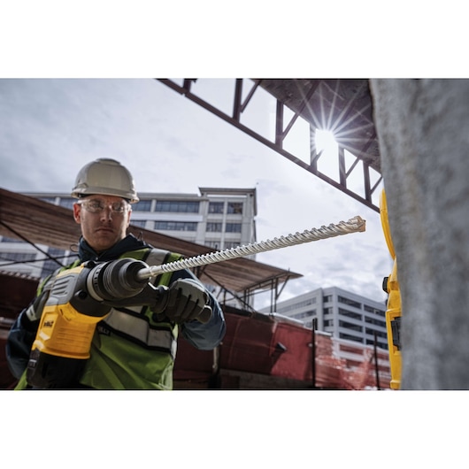 Worker uses a DEWALT drill with DT60825 bit at a construction site with buildings in the background.