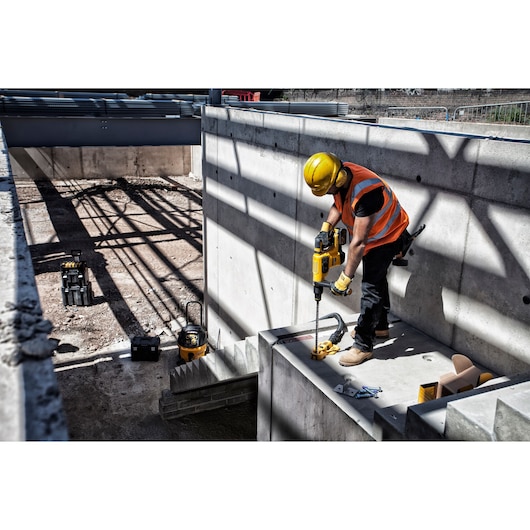 Worker uses DEWALT power tool to drill concrete at construction site, wearing safety gear and orange vest.