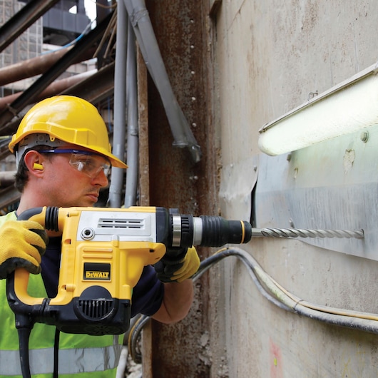 Worker uses DEWALT drill DT60825 for drilling into concrete wall at construction site, wearing safety gear.
