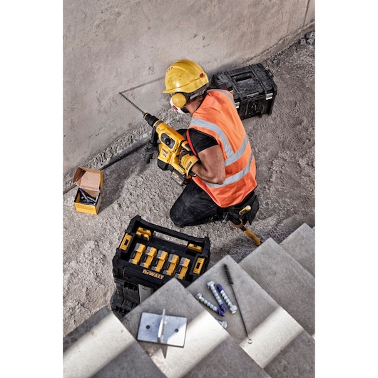 Worker uses DEWALT drill for concrete with toolboxes and hardware nearby on a construction site.