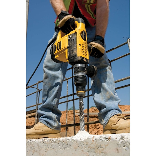 Person drilling concrete with a yellow DEWALT tool at a construction site, wearing gloves and work boots.