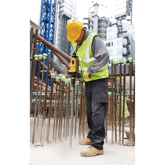 Worker using a DEWALT drill at construction site. Wearing safety gear and surrounded by rebar and buildings.