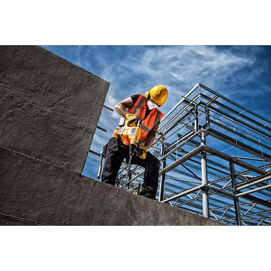 Worker uses DEWALT drill on concrete at construction site, wearing helmet and vest under a blue sky.
