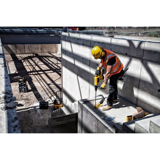 Worker using DEWALT drill on concrete at construction site, wearing safety gear and orange vest.