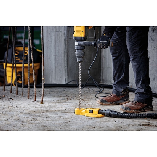 Person using a DEWALT hammer drill with dust extraction on concrete at a construction site. Rebar visible in background.