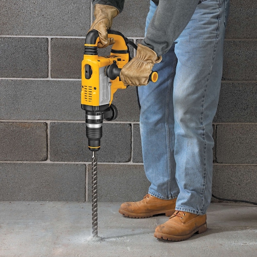 Person wearing gloves drills into concrete floor using a yellow DEWALT rotary hammer with long bit.