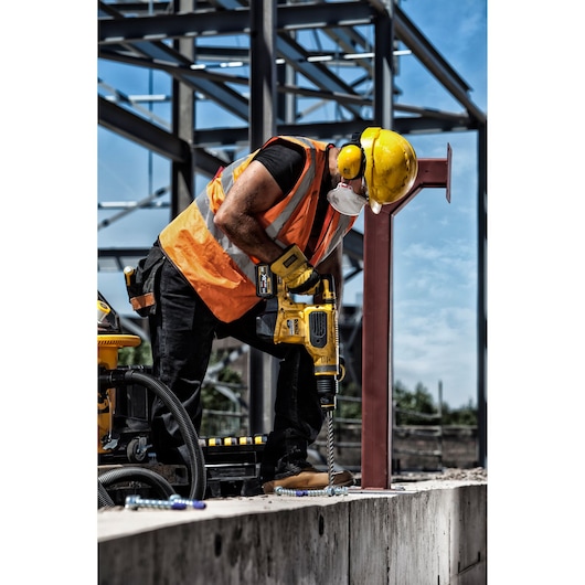 Worker uses a DEWALT drill on concrete at a construction site, wearing safety gear and orange vest.