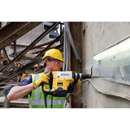 Person uses a DEWALT rotary hammer drill on a concrete wall at a construction site, wearing safety gear.