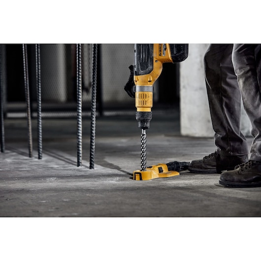 Person using DEWALT hammer drill with drill bit on concrete floor at construction site.